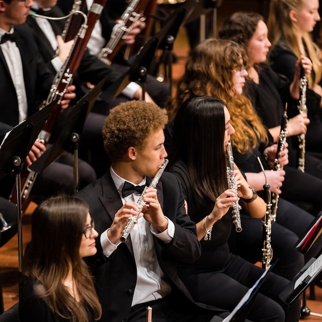 Male student playing a flute with other students playing woodwind instruments around him in a concert