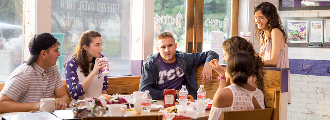 Off campus, a group of six TCU students laugh and eat hamburgers together at a large table at Dutch's burgers.