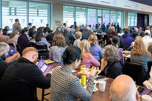 A group of faculty and staff gather at tables in the Legends Club.