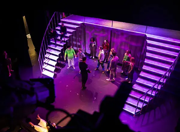 Theatre students rehearse on a set with wraparound stairs, flooded in purple lights